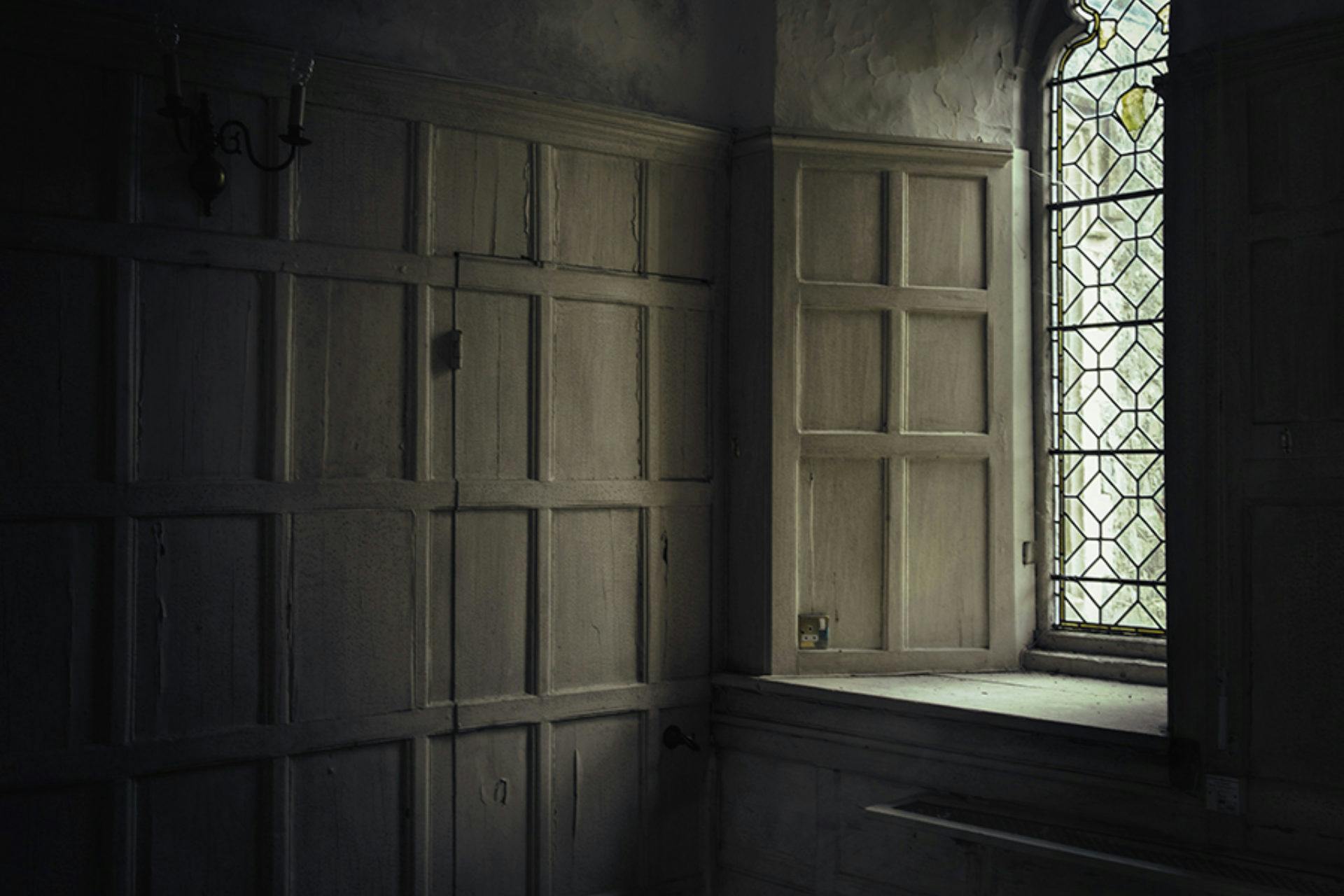 Dimly lit room with vintage wood panelling and a leaded window letting in minimal light, creating a moody atmosphere.