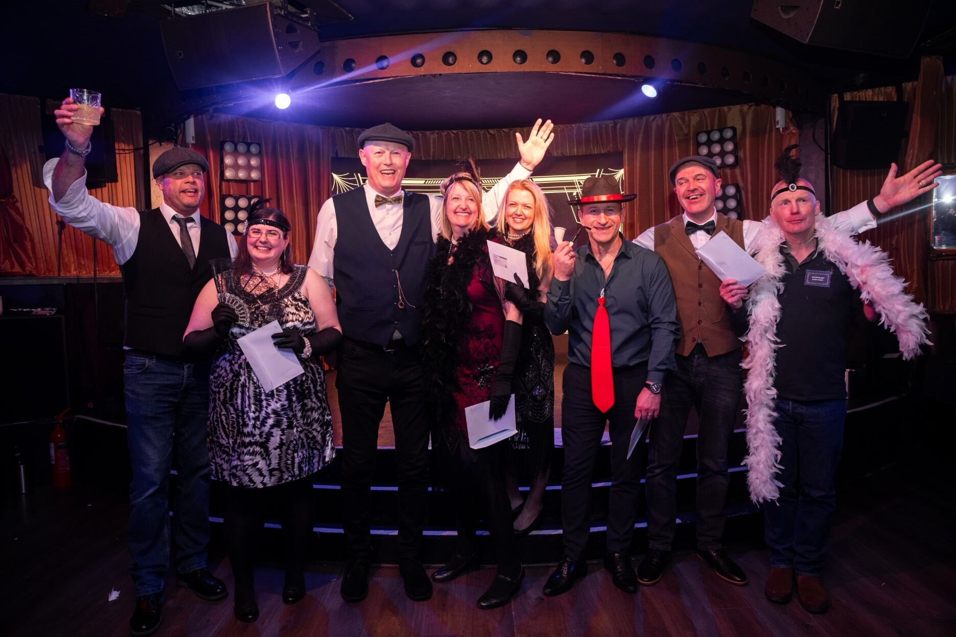 Group of people dressed in themed costumes, smiling and posing on a stage at an event, with some holding papers and raising their hands.