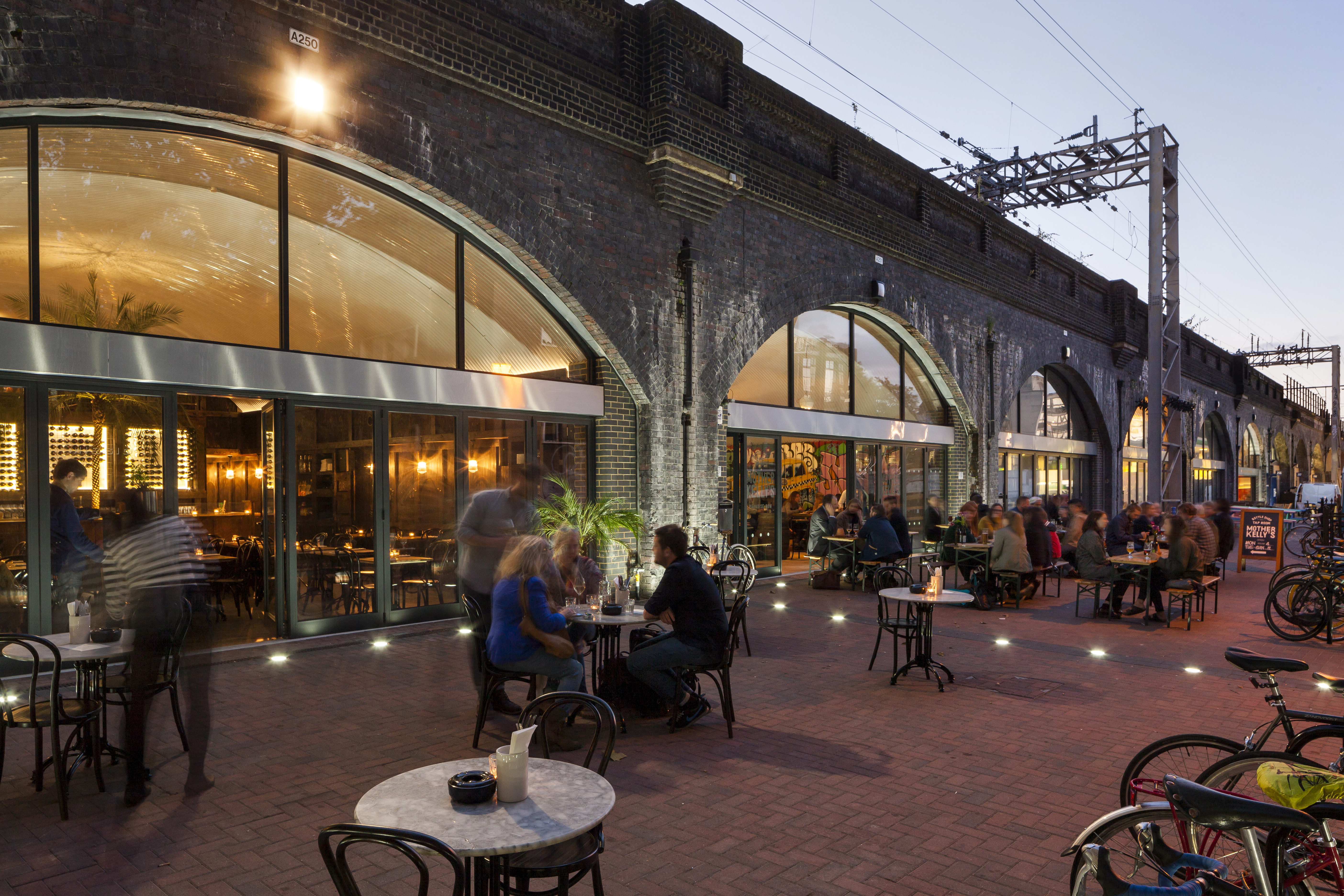Outdoor dining area in front of arched, glass-fronted restaurant spaces beneath a brick railway viaduct, with people seated at tables in the evening