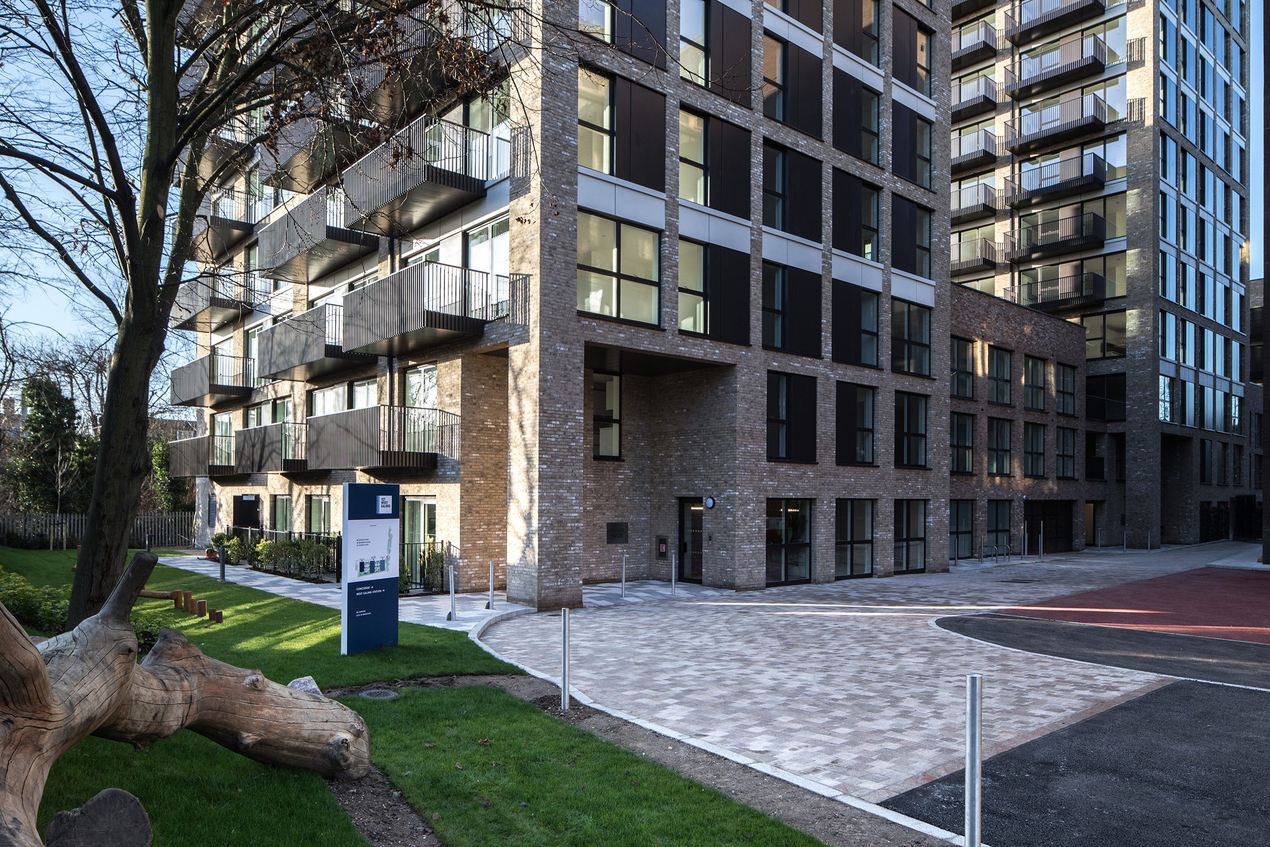 Modern apartment building with brick facade and balconies, fronted by a paved courtyard and green landscaping.