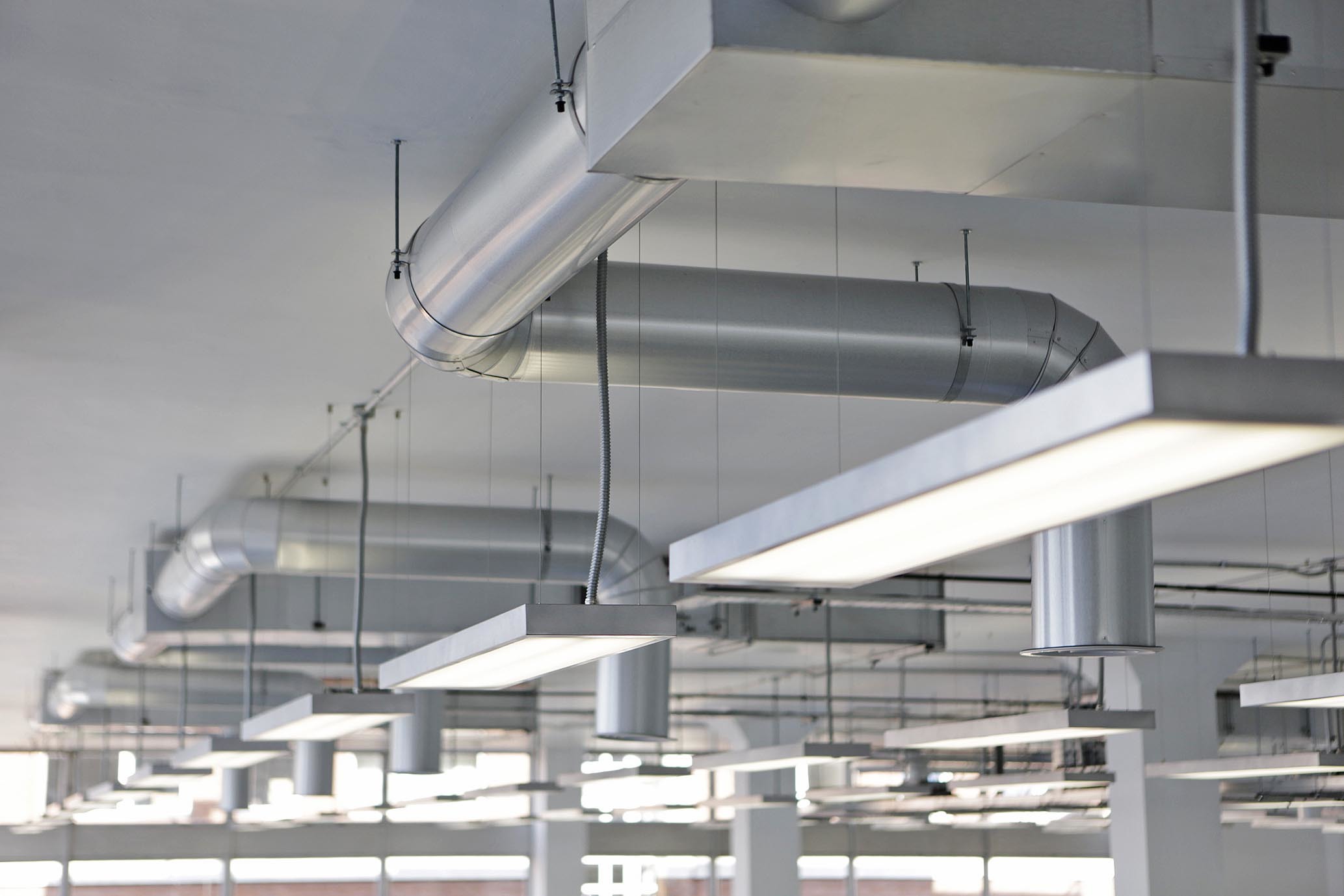 Industrial ceiling with exposed silver ventilation ducts, white rectangular light fixtures, and grey structural beams in large interior space.