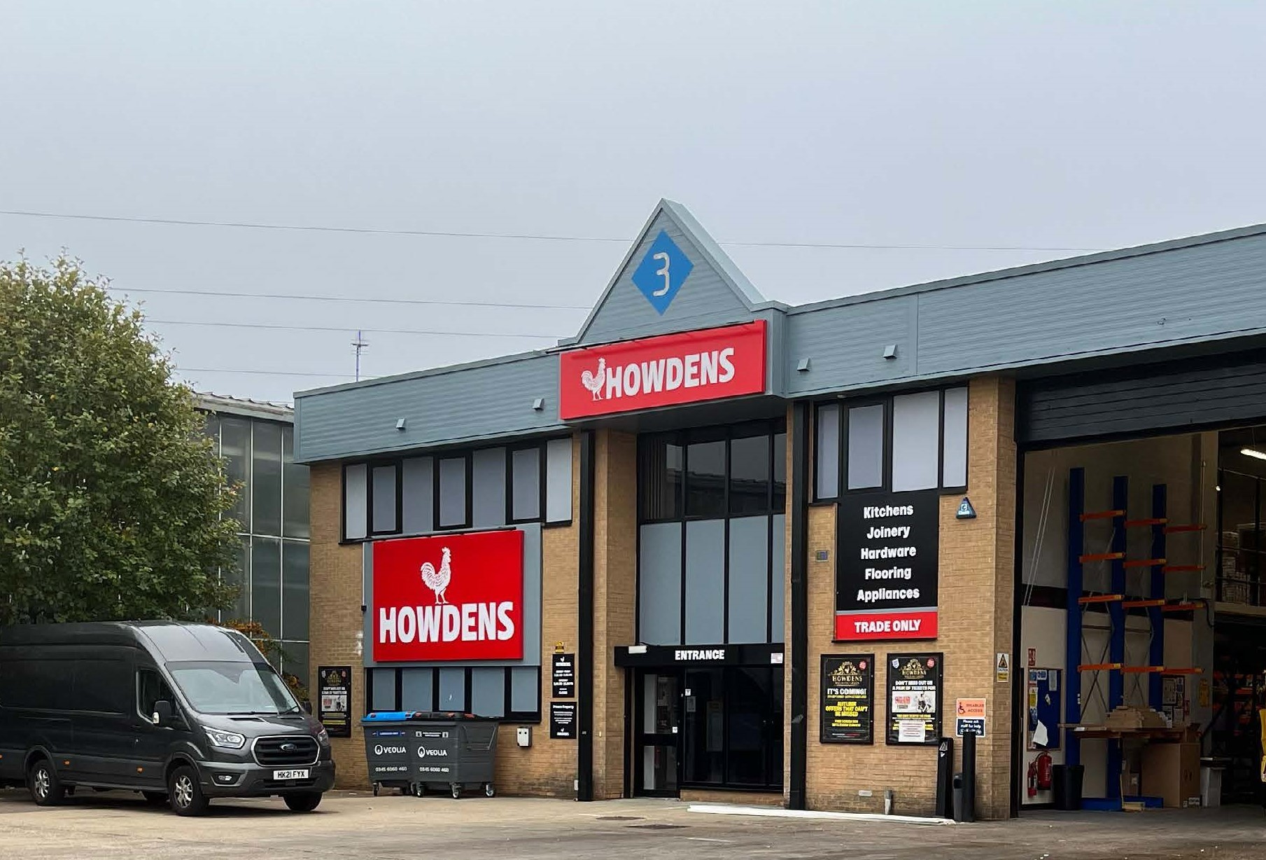 Exterior of a Howdens trade store, with a red sign and large windows, and a black van parked outside