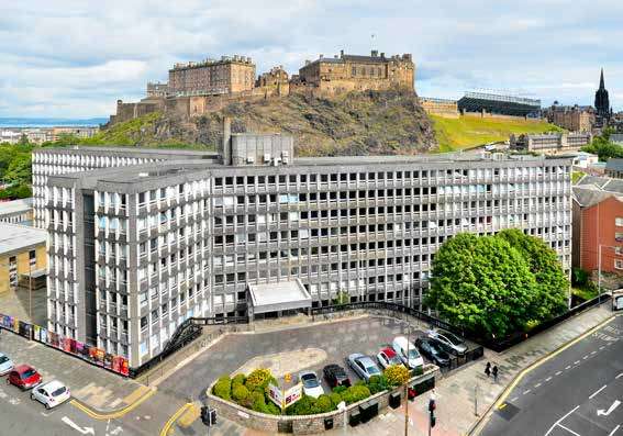 Modern office building in the foreground with a historic castle on a hill in the background, under a partly cloudy sky.