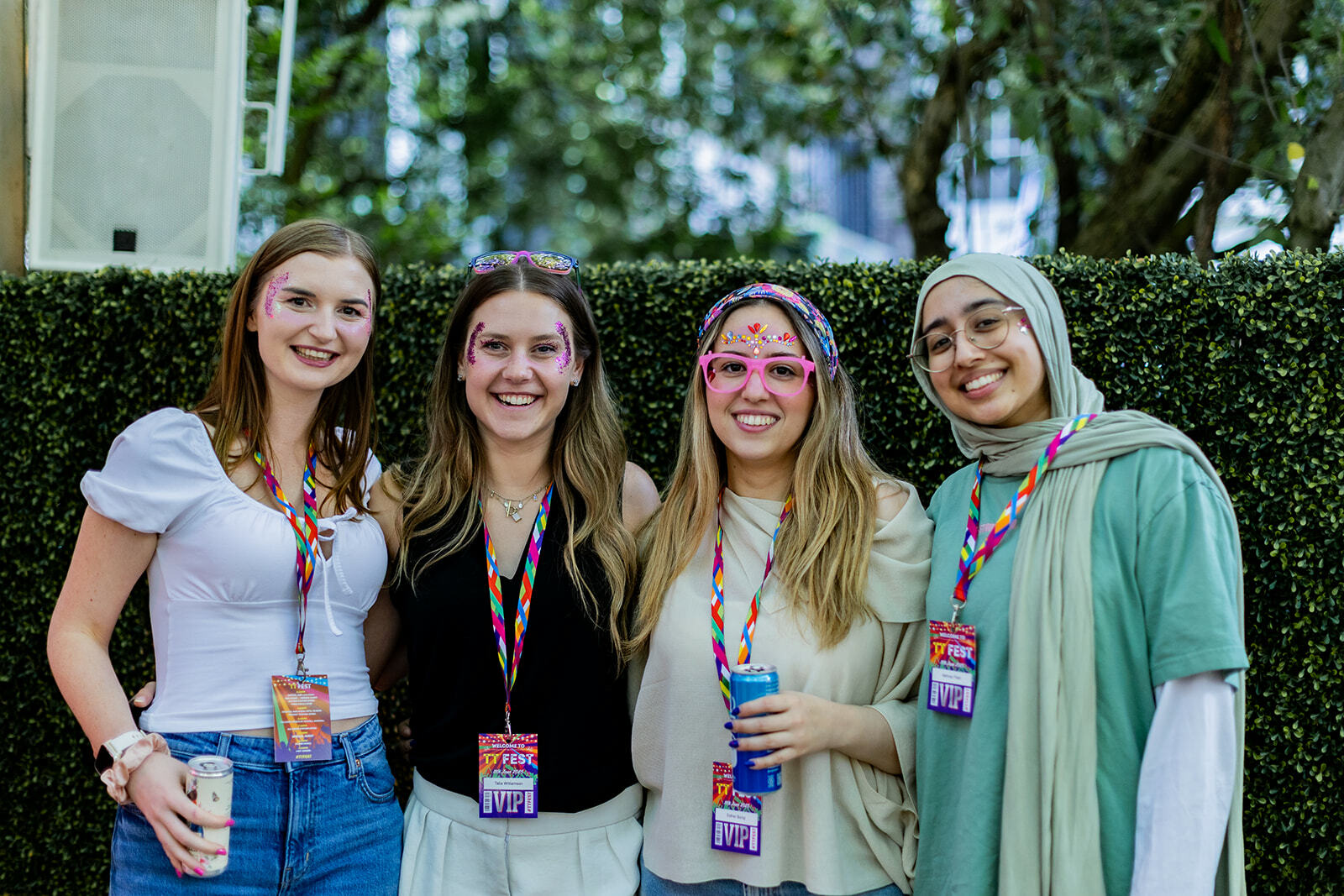 Four young women wearing lanyards stand together smiling outdoors, with green hedge and trees in background.