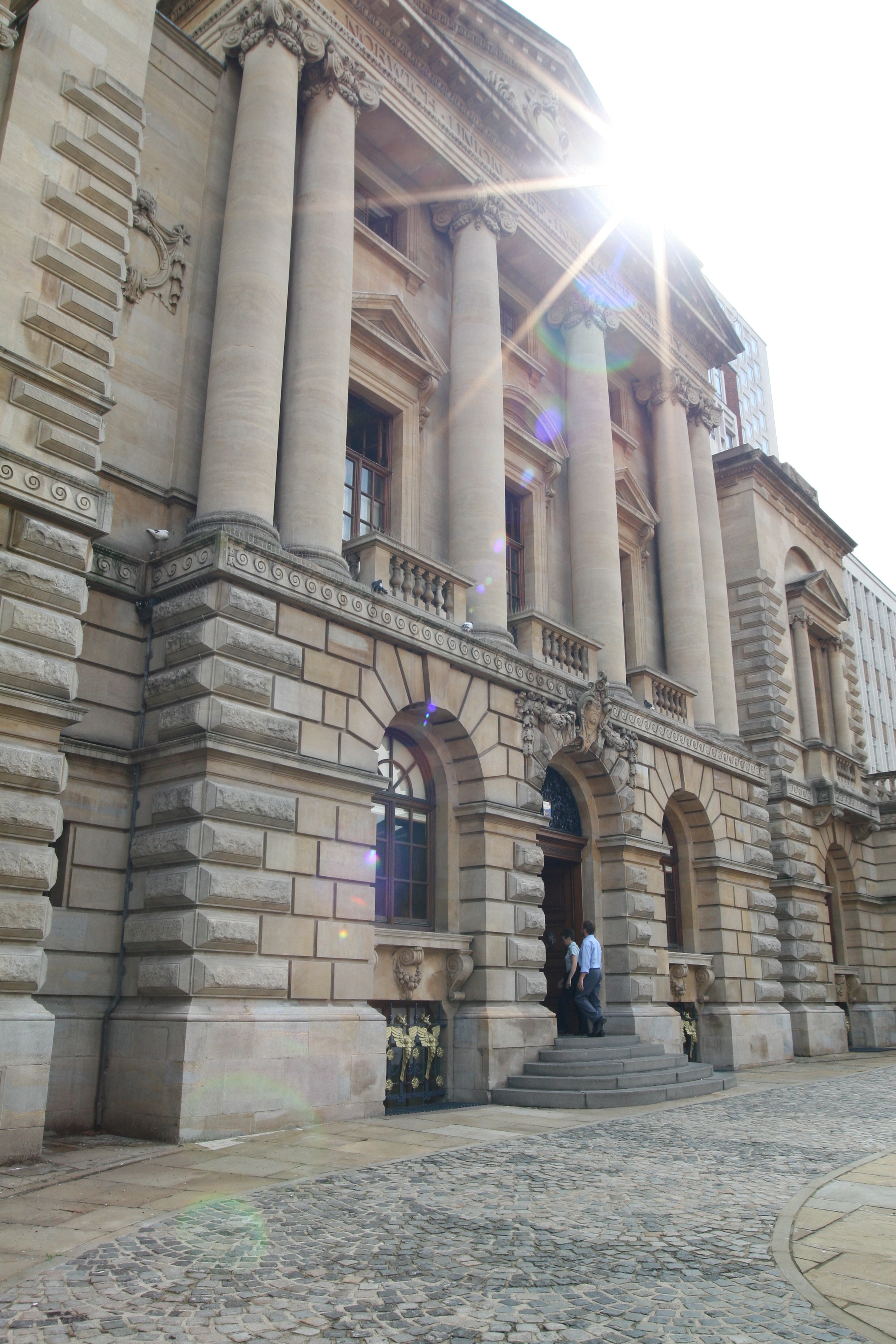 Sunlit exterior of a grand stone building with tall columns and arched entrances, featuring cobblestone paving in the foreground