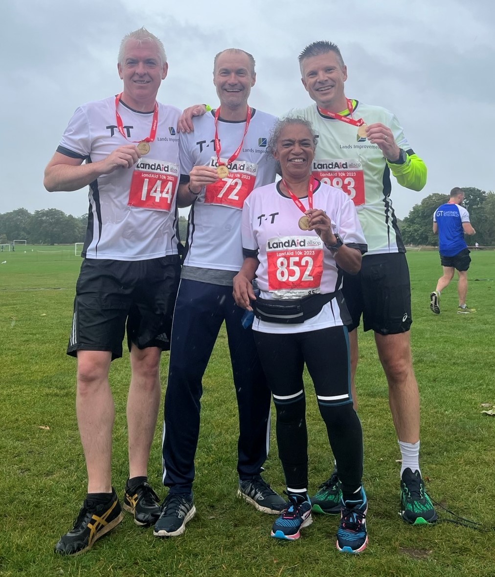 Four runners wearing race bibs and medals, standing together and smiling after completing a race on a grassy field.