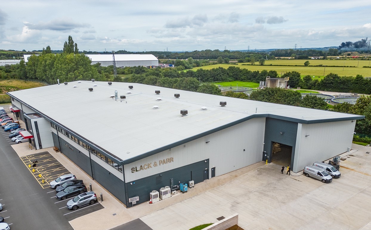Aerial view of a large industrial building labeled 'Slack & Parr,' with parking spaces and surrounding fields in the background