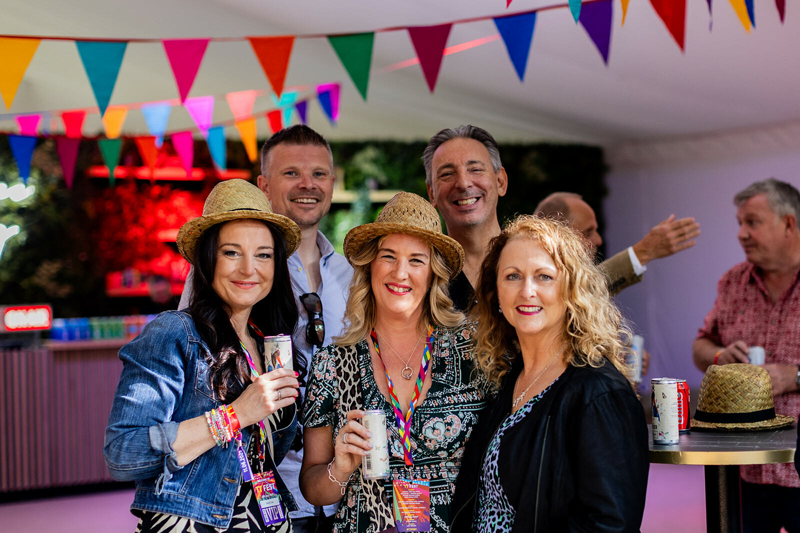 Group of adults at party holding drinks under colourful triangular bunting, smiling at camera in indoor venue.