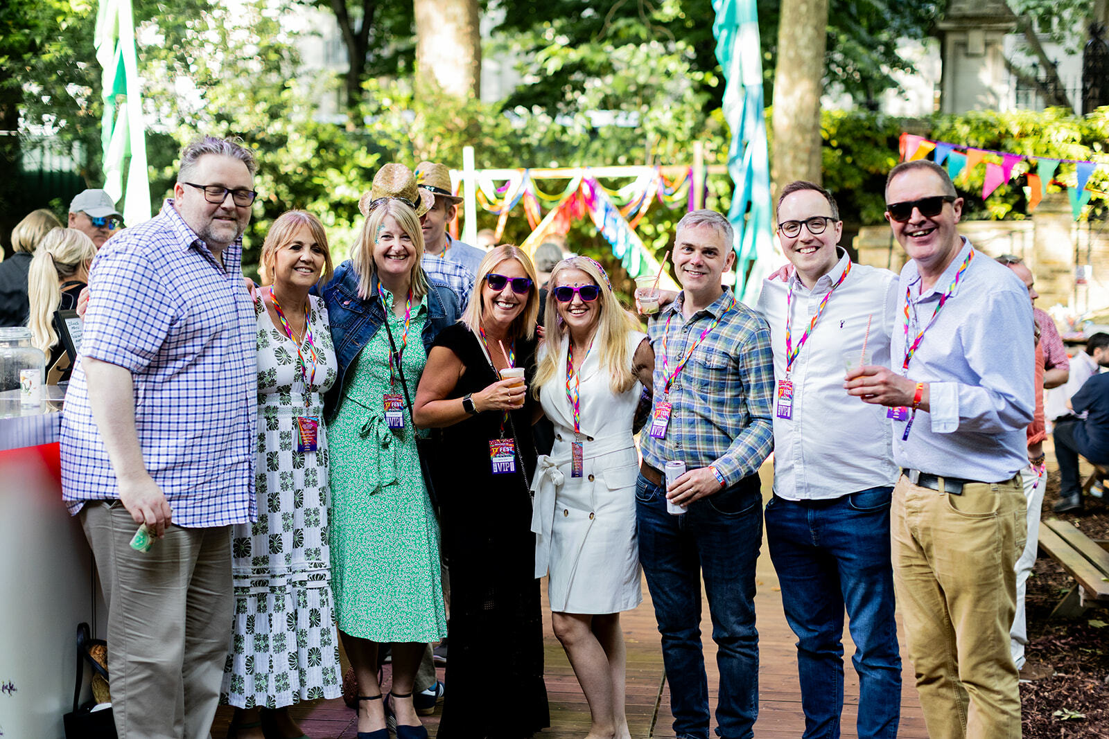 Group of eight people posing together at outdoor event with colourful bunting and trees in background, wearing casual summer clothing.