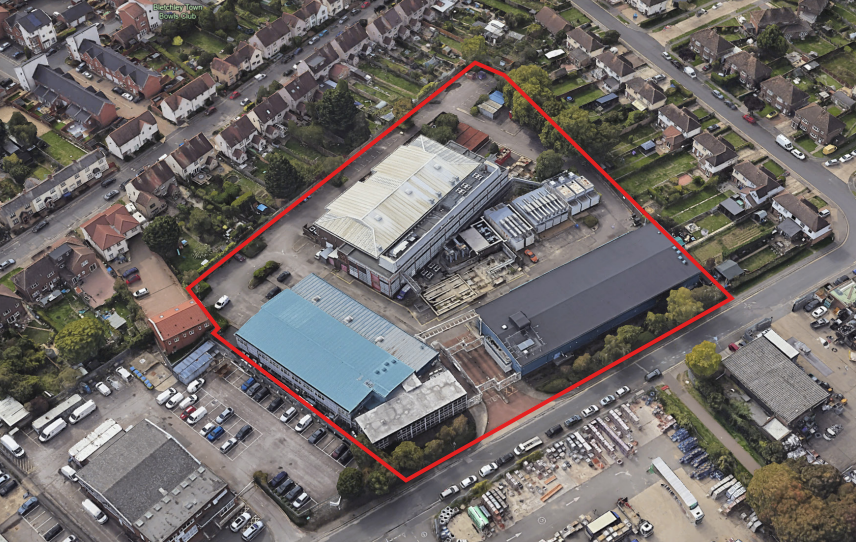 Aerial view of an industrial complex outlined in red. The site includes multiple large buildings with metal roofs, surrounded by residential neighborhoods, roads, and parked vehicles.