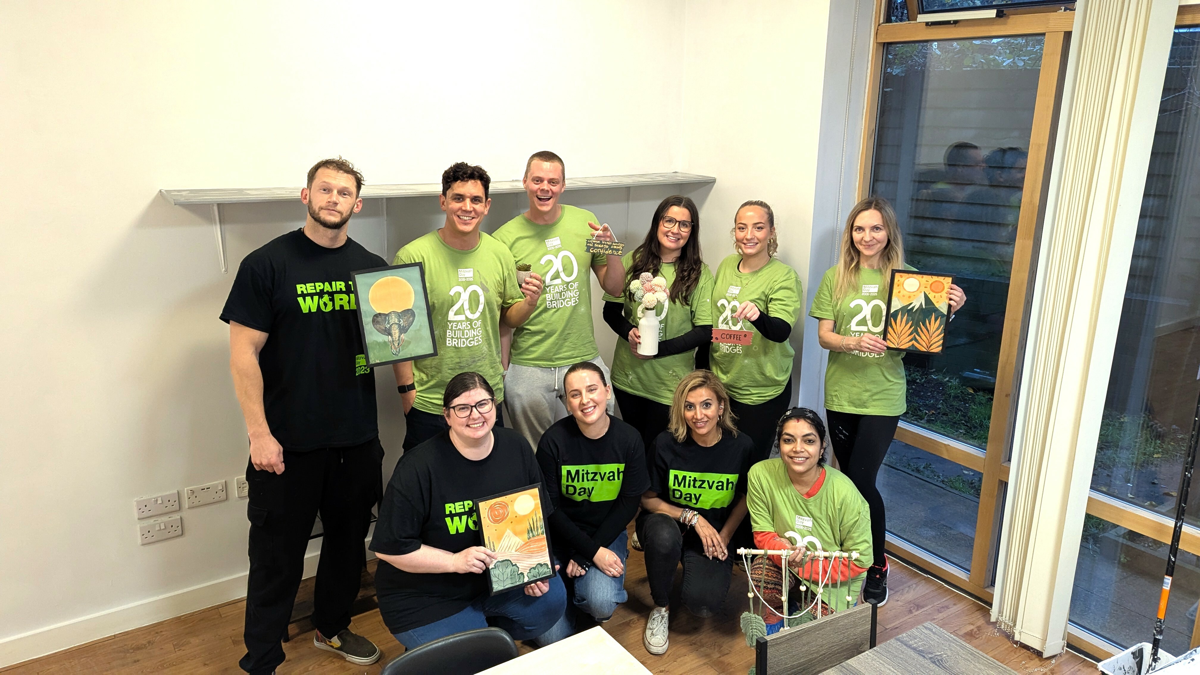 Ten people in green shirts and black clothing pose indoors holding certificates, with windows visible on right side of white-walled room.