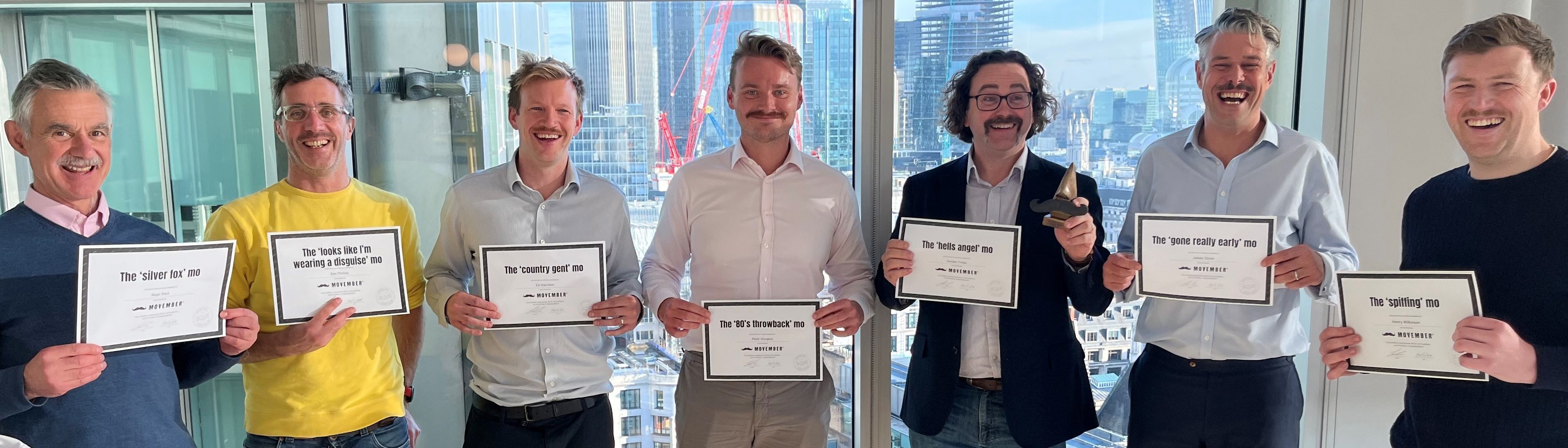 Group of seven men smiling and holding certificates, standing in front of a large window with a cityscape in the background