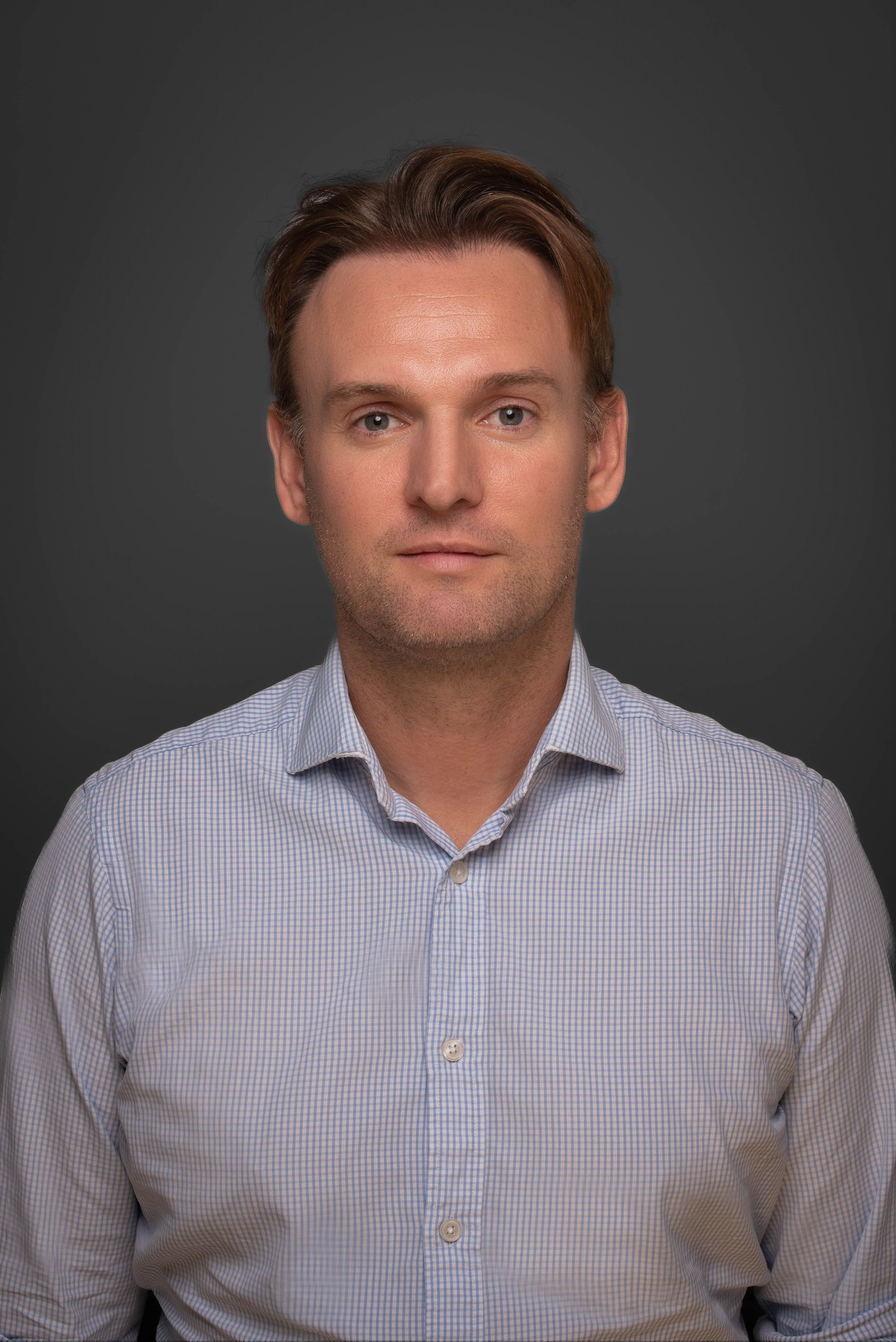 Professional headshot of man with brown hair wearing light blue striped shirt against grey background.