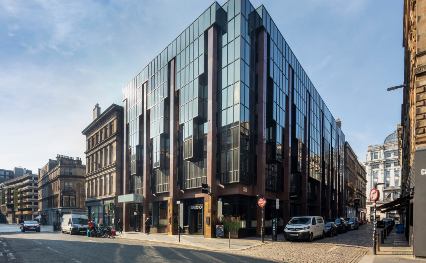 Modern dark glass building with vertical metal strips on cobbled street, flanked by traditional Victorian architecture.