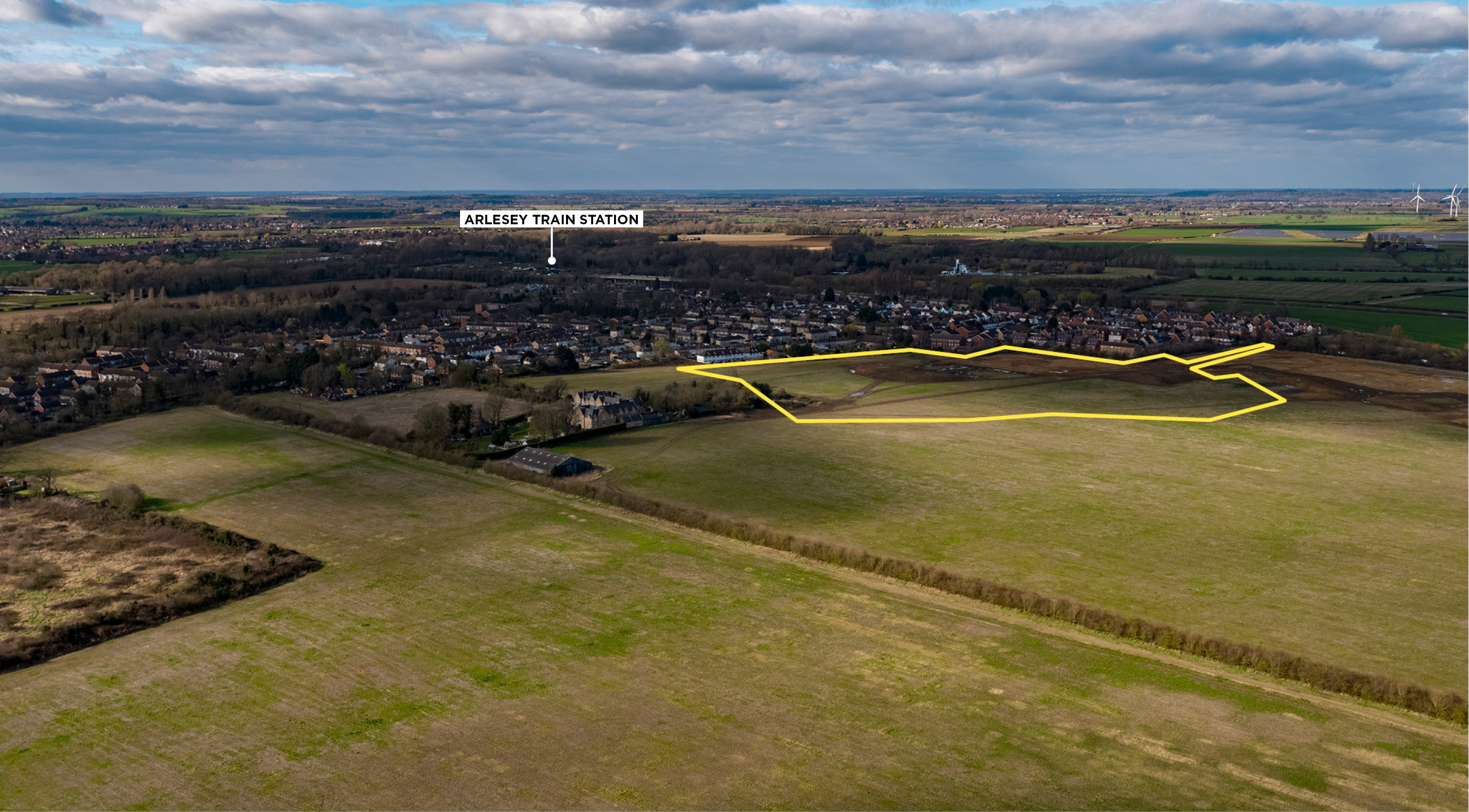 Aerial view of a rural landscape with open fields and a small town in the distance. A section of land is outlined in yellow near Arlesey Train Station.