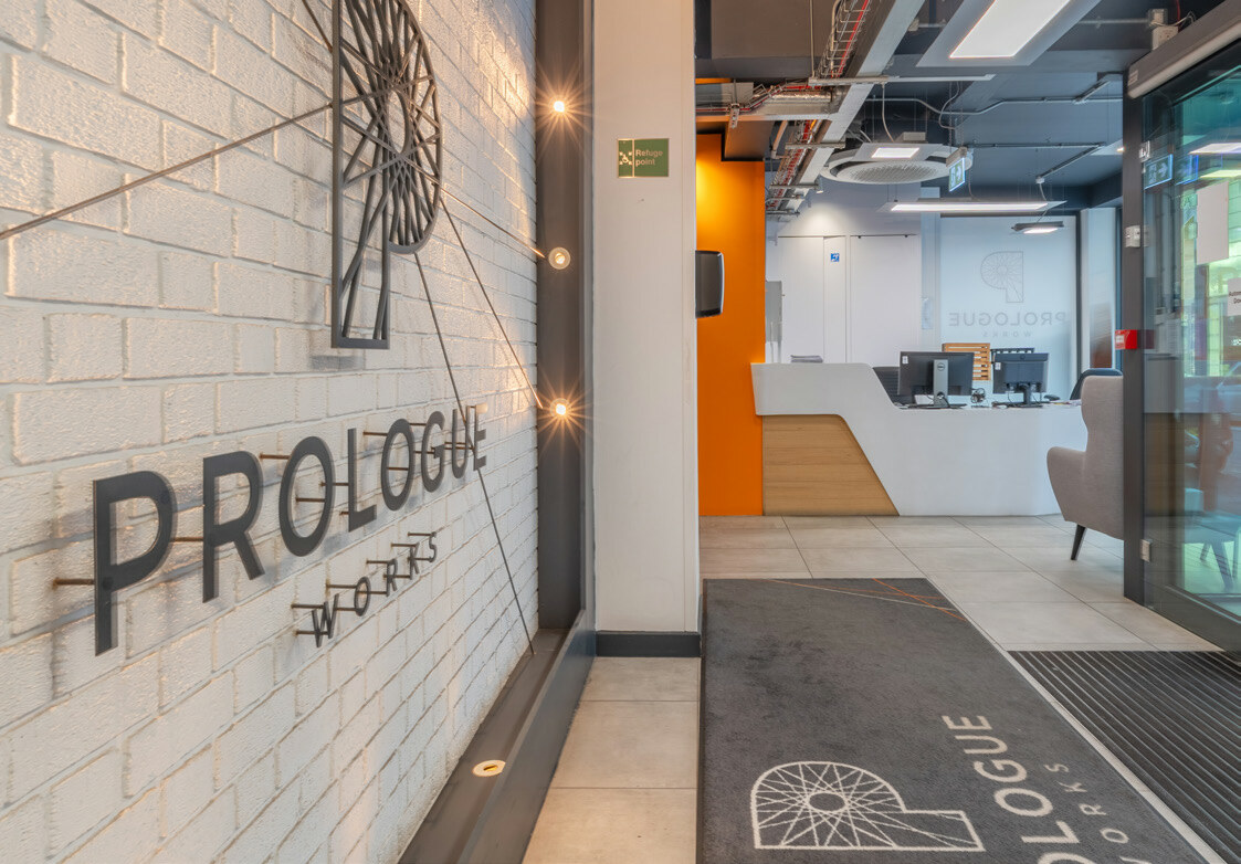 Modern office entrance with white brick wall featuring "PROLOGUE" text and circular wire art, orange accent wall, glass doors and reception area.