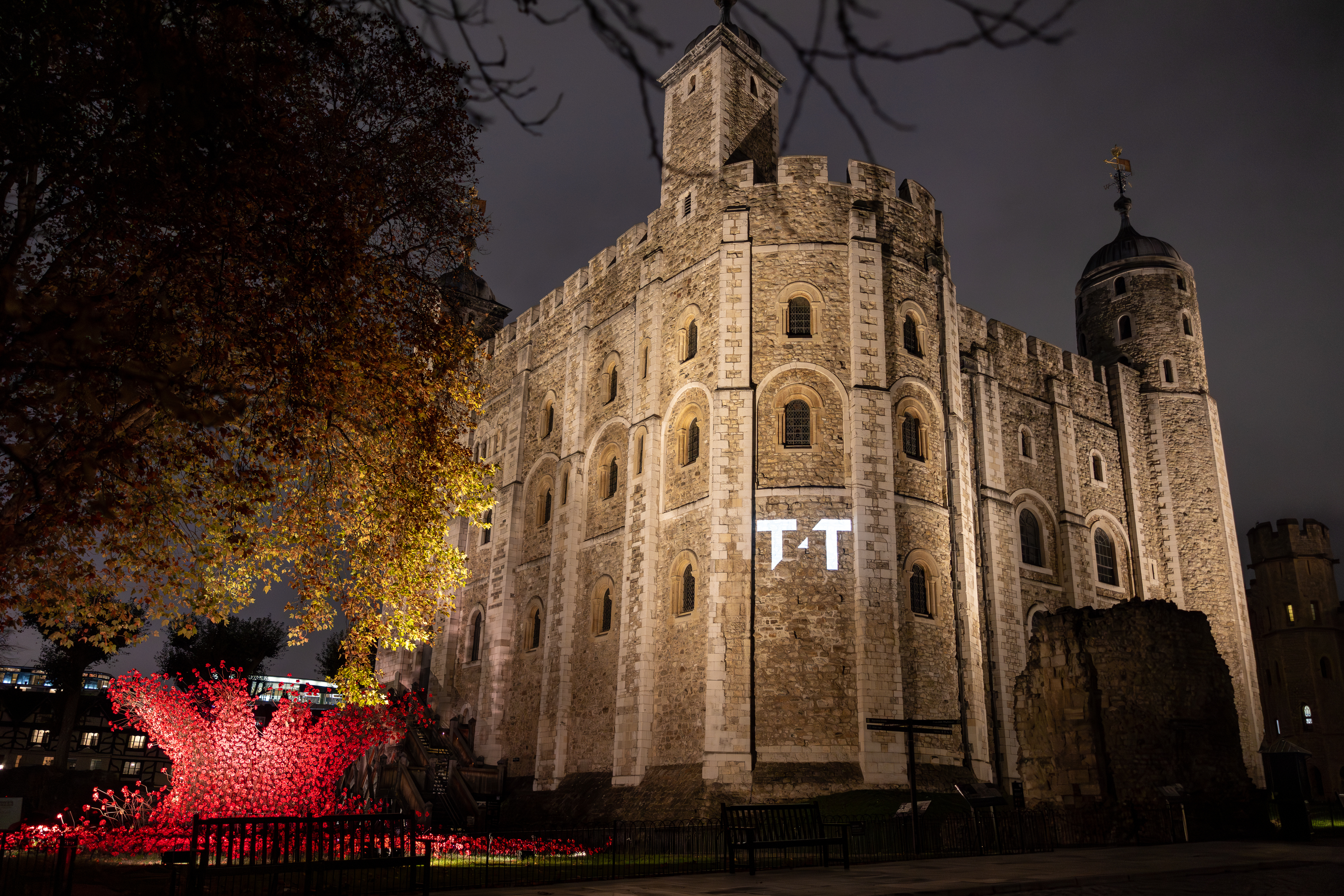 Tower of London illuminated at night with warm golden lighting on stone walls, red poppies installation visible, dark sky background.
