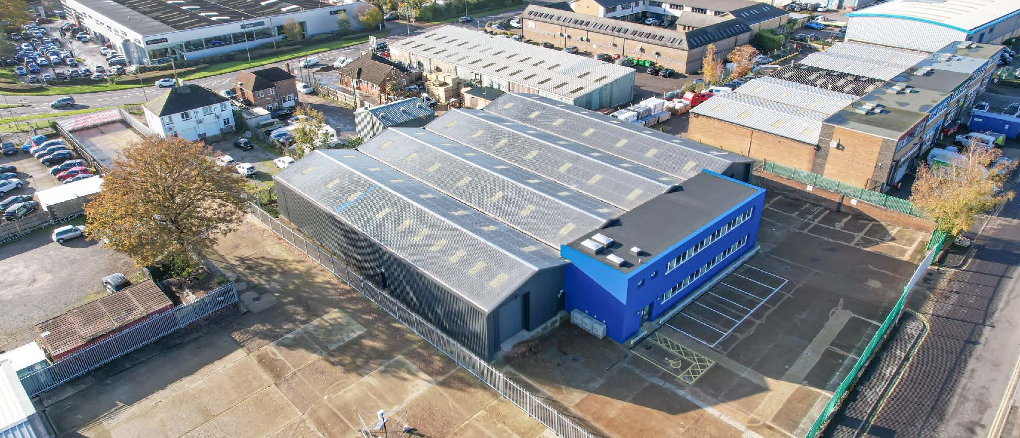Aerial view of a large industrial warehouse with a blue office section, located in a commercial area with surrounding buildings and parking spaces.