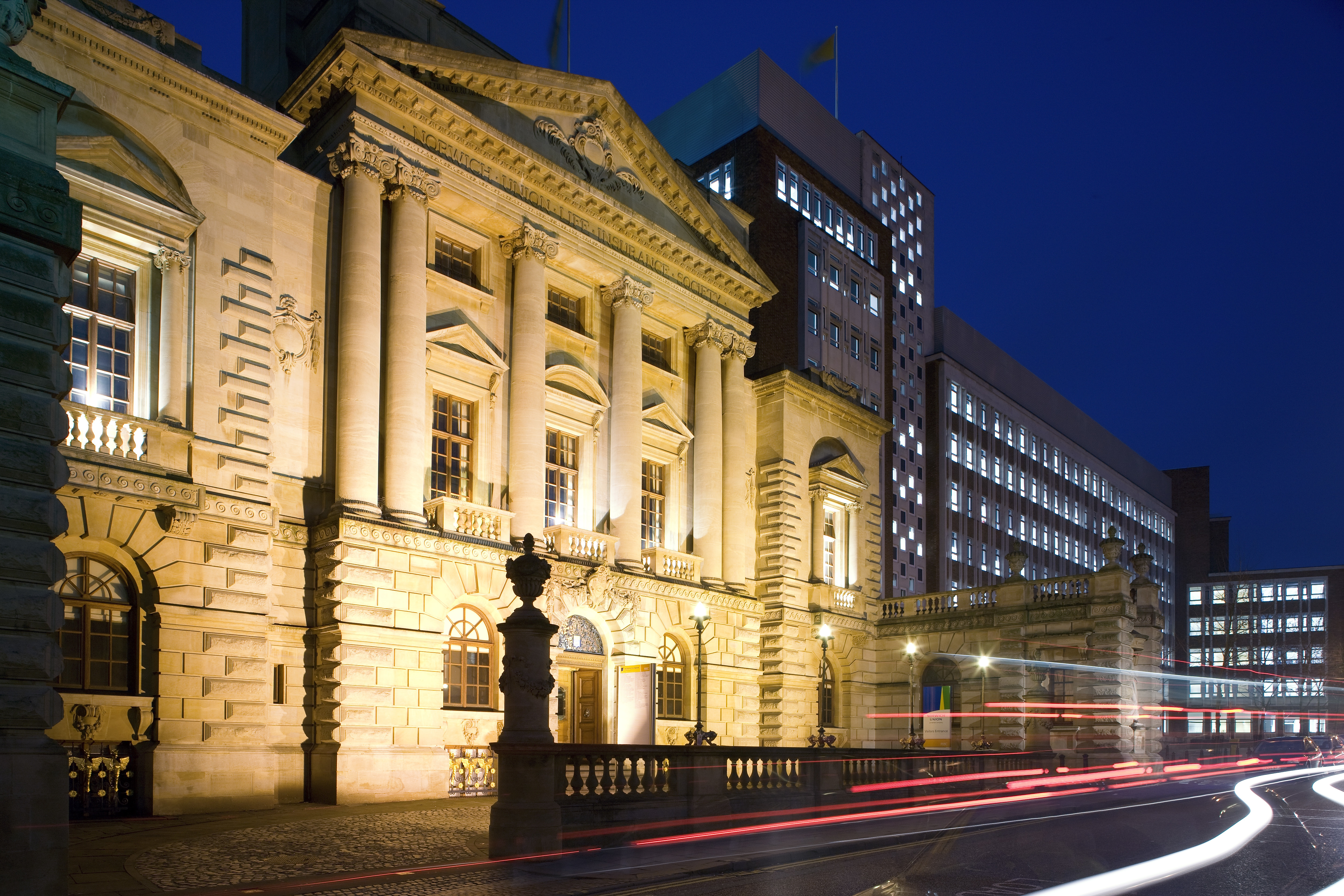 Historic building illuminated at night with classical architecture, next to a modern building, with light trails from passing traffic.