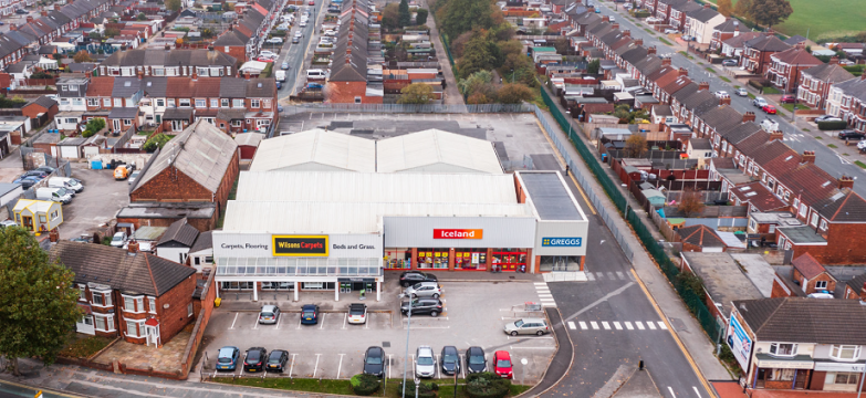 Aerial view of a retail complex featuring stores such as Iceland and Greggs, with a parking lot in front, situated in a residential neighborhood