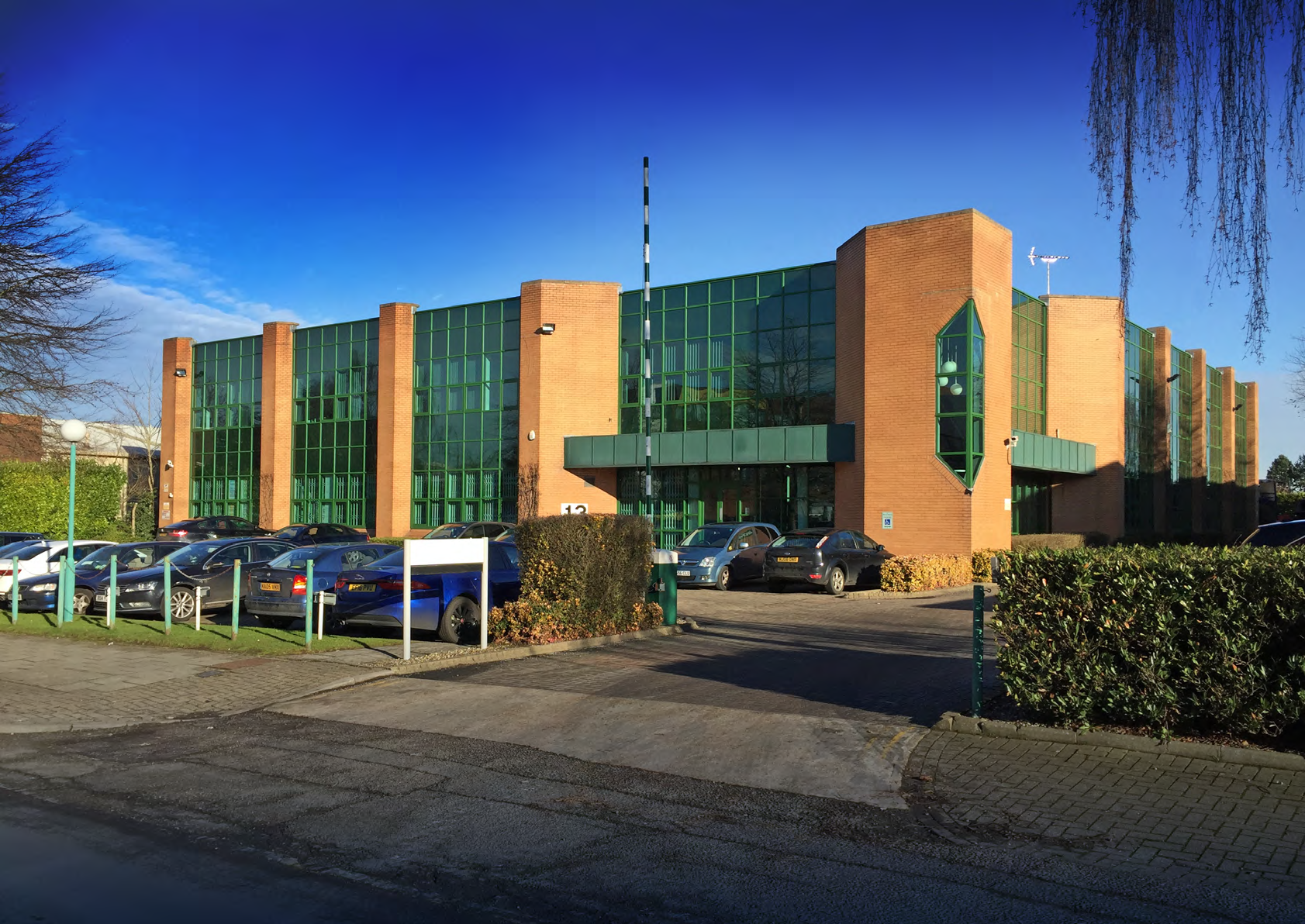 Exterior view of a modern, two-story brick office building with large green-tinted windows and a parking lot in front.