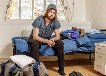 A man with a beard and beanie hat is sitting on the edge of a bed