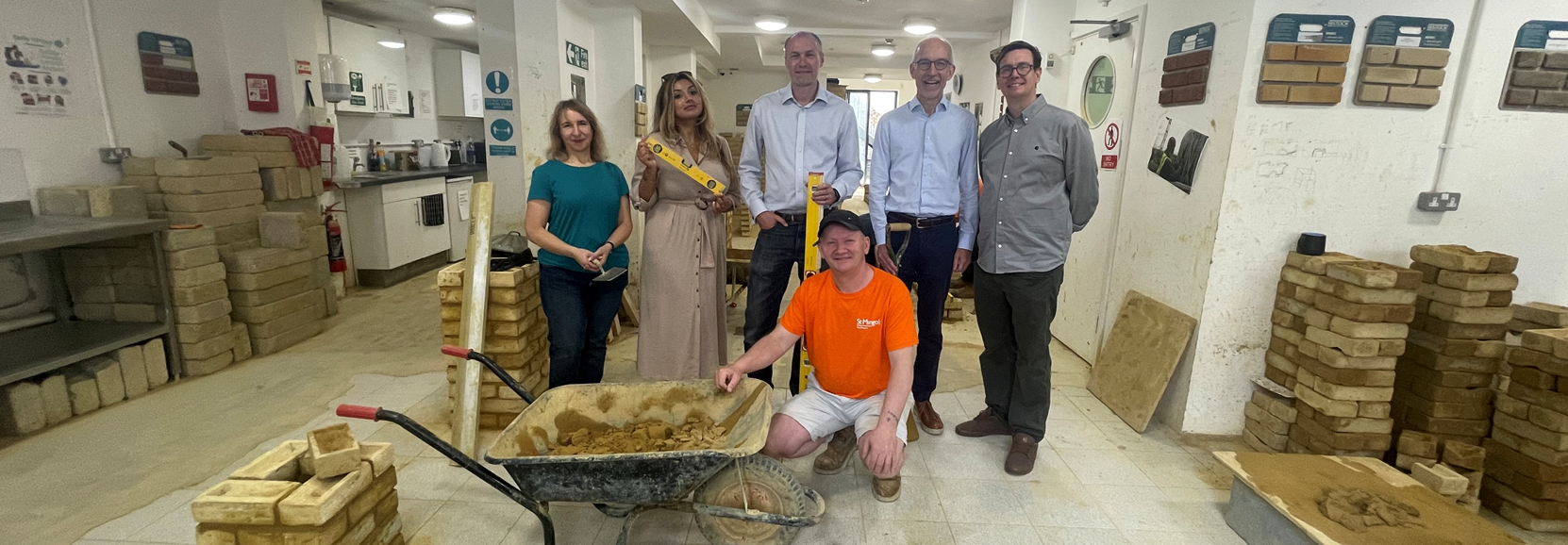 Group of six people standing indoors in front of stacked bricks, with a person kneeling in front holding a wheelbarrow filled with building materials.