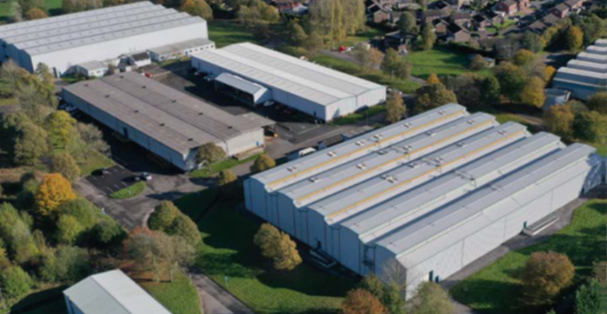 Aerial view of a large industrial complex with several warehouse buildings, surrounded by trees and greenery in a suburban area.
