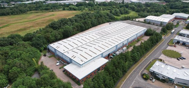 Aerial view of a large industrial warehouse surrounded by trees, located in a commercial area with nearby roads and buildings.
