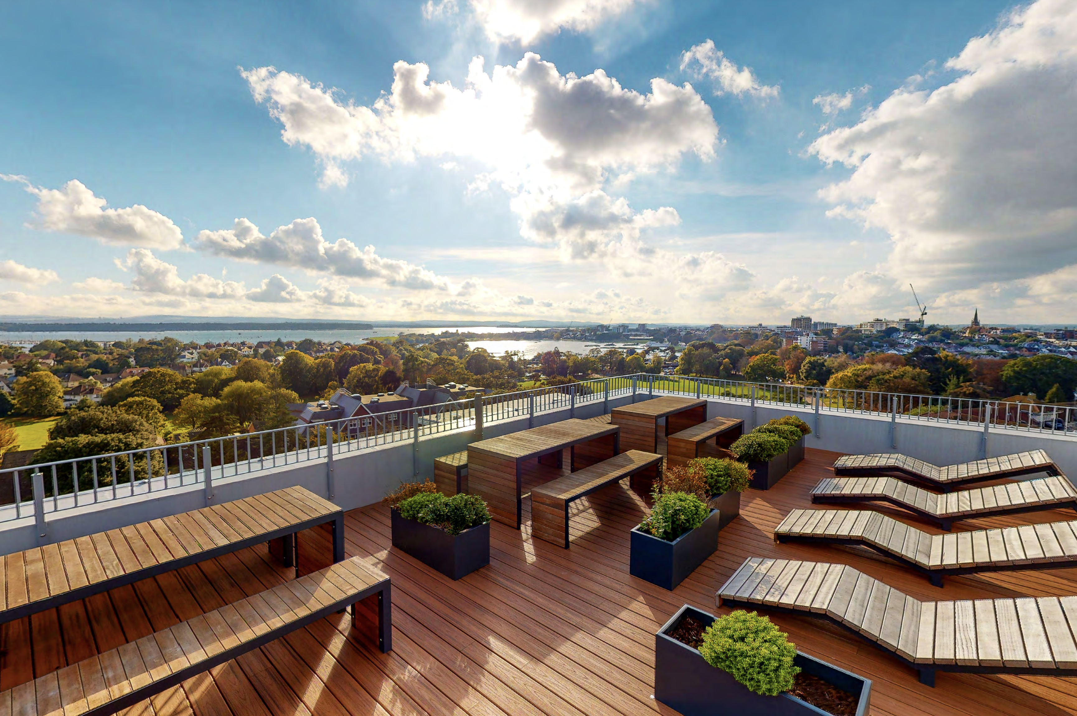 A rooftop terrace with wooden decking, featuring several sun loungers, benches, and potted plants. The terrace overlooks a scenic view of trees, houses, and a body of water in the distance under a partly cloudy sky with sunlight breaking through.
